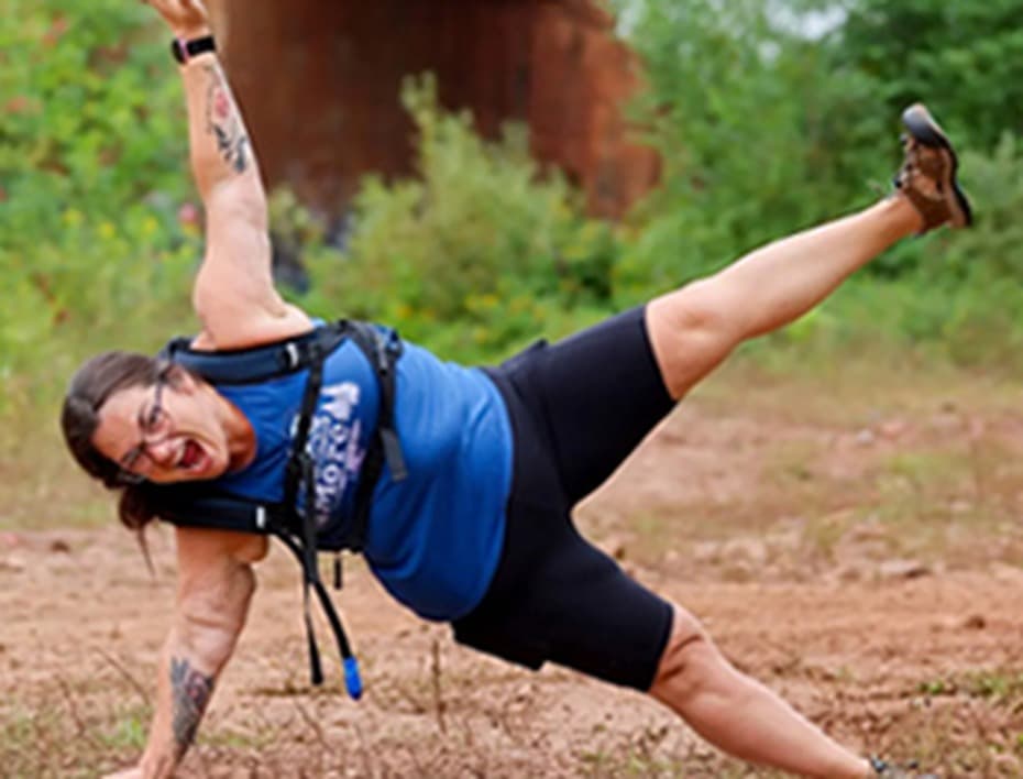 Chrissy holding a side plank on a trail, one leg raised, laughing